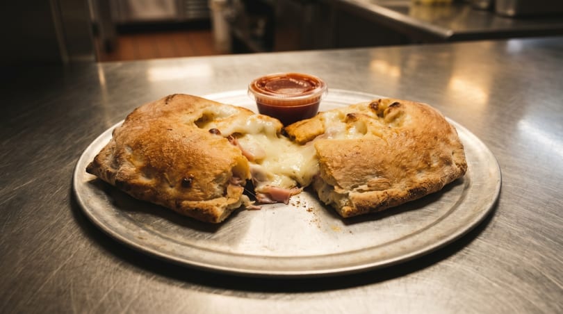 A golden-brown calzone, split open to reveal melted cheese and ham, served with a side of marinara sauce.