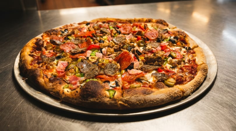 A large, fully loaded pizza with various meats, vegetables, and cheese on a metal tray.