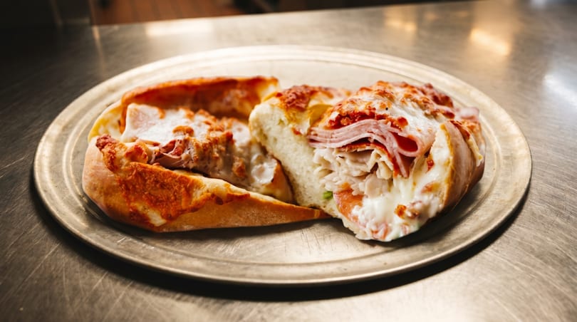 Two halves of a cheesy calzone filled with ham and other meats on a metal tray.
